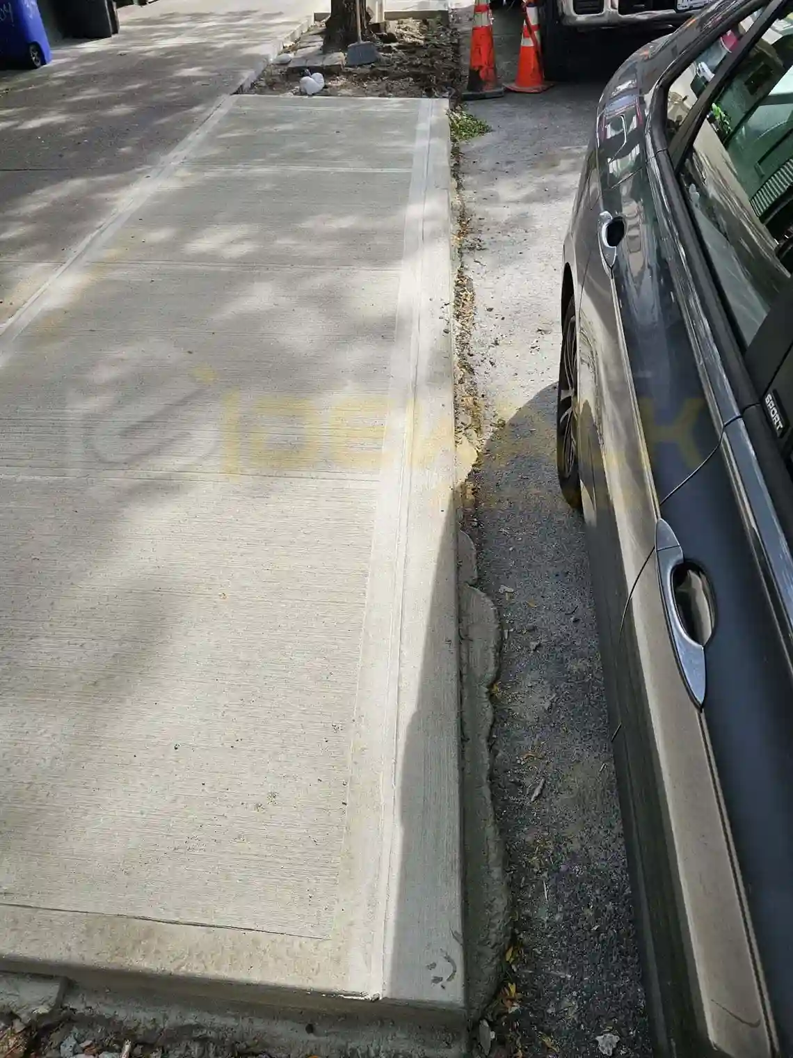A cracked concrete sidewalk next to a curbside drain grate with some grass growing in the cracks.